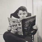 Boy sitting in chair, one foot over the other knee, reading a superman comic book