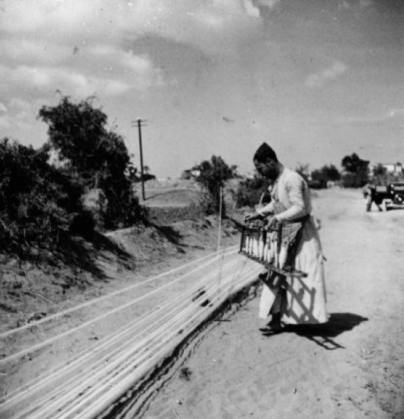 The man is standing outside next to many long rows of some kind of thread, and he appears to be weaving in more threads at a 90 degree angle to the long ones.