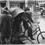 Chinese youth on a bicycle passenger cart in the rain by the side of road, perhaps in the company of a couple others with similar rigs because one person and other wheels are visible; shops and and truck in background.