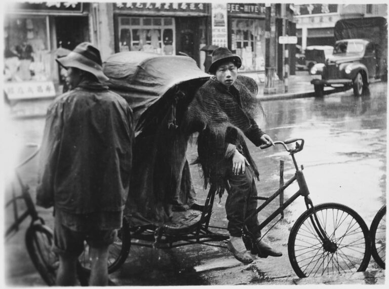 Chinese youth on a bicycle passenger cart in the rain by the side of road, perhaps in the company of a couple others with similar rigs because one person and other wheels are visible; shops and and truck in background.