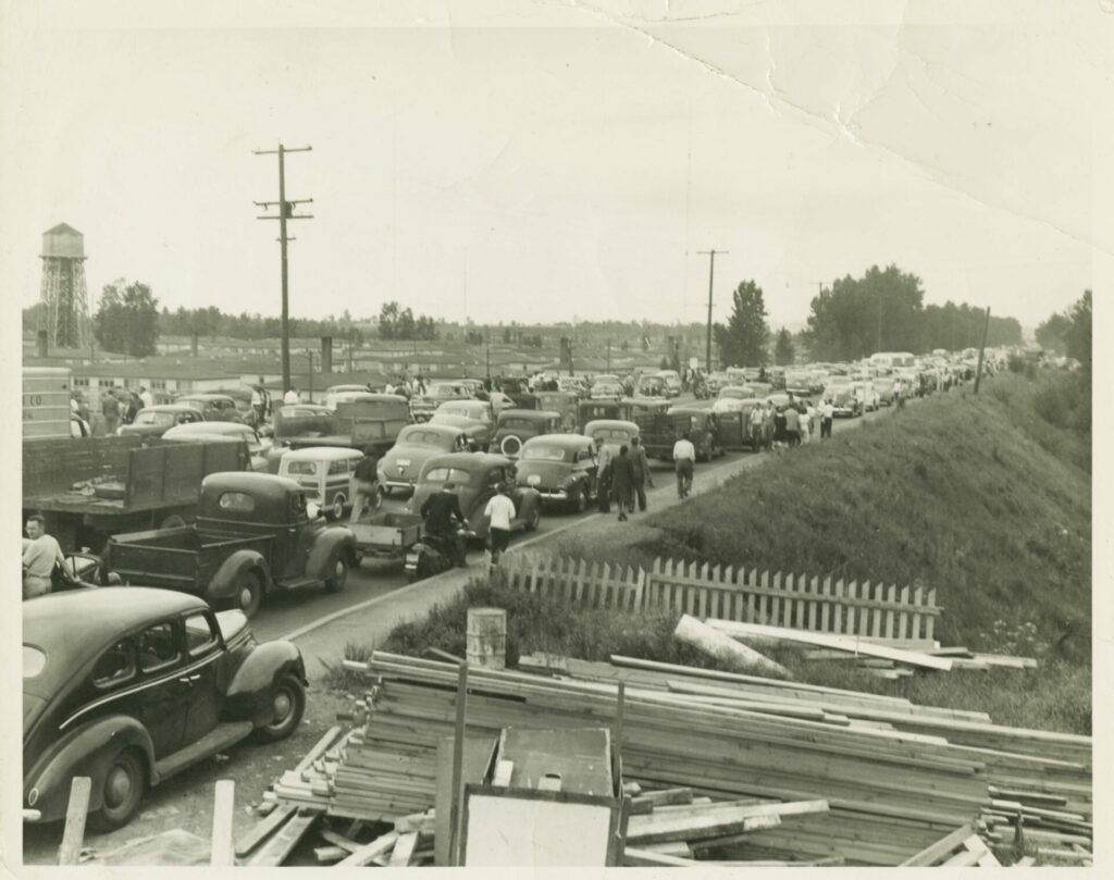 A line of cars parked on Denver Avenue in Vanport, Oregon. The drivers abandoned their cars on the road and fled on foot after a traffic jam blocked their exits. Pedestrians are visible on the road attempting to escape the rising flood waters during the Vanport flood.
