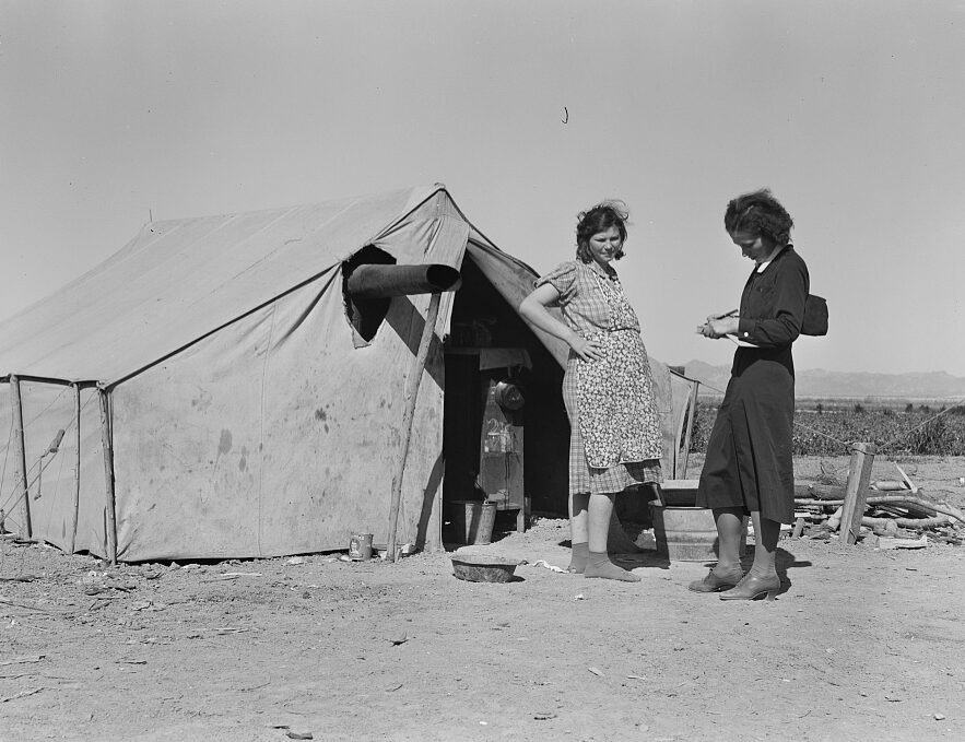 Female interviewer stands before pregnant woman before a tent in a desert setting. Interviewer writes notes in her notebook.