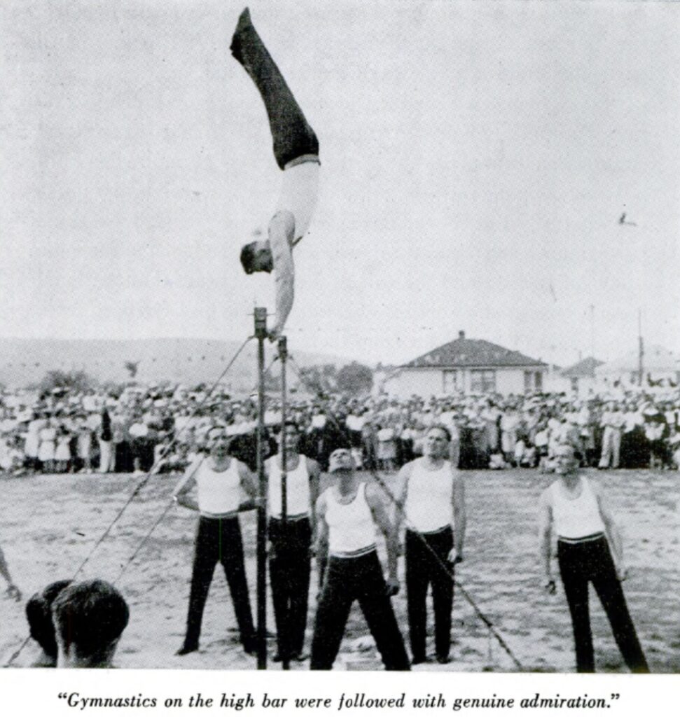 Black and white photo with male gymnasts. One gymnast is performing a handstand on the high bar while 5 other gymnasts stand below. A large crowd is gathered in the background in front of workers' simple, small white homes. In-image caption reads: "Gymnastics on the high bar were followed with genuine admiration."