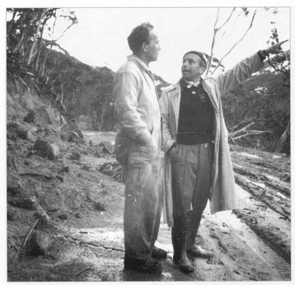 Two mean stand in a muddy clearcut area being used as a road. One points up with his left hand. They are wearing workclothes. Black and white.