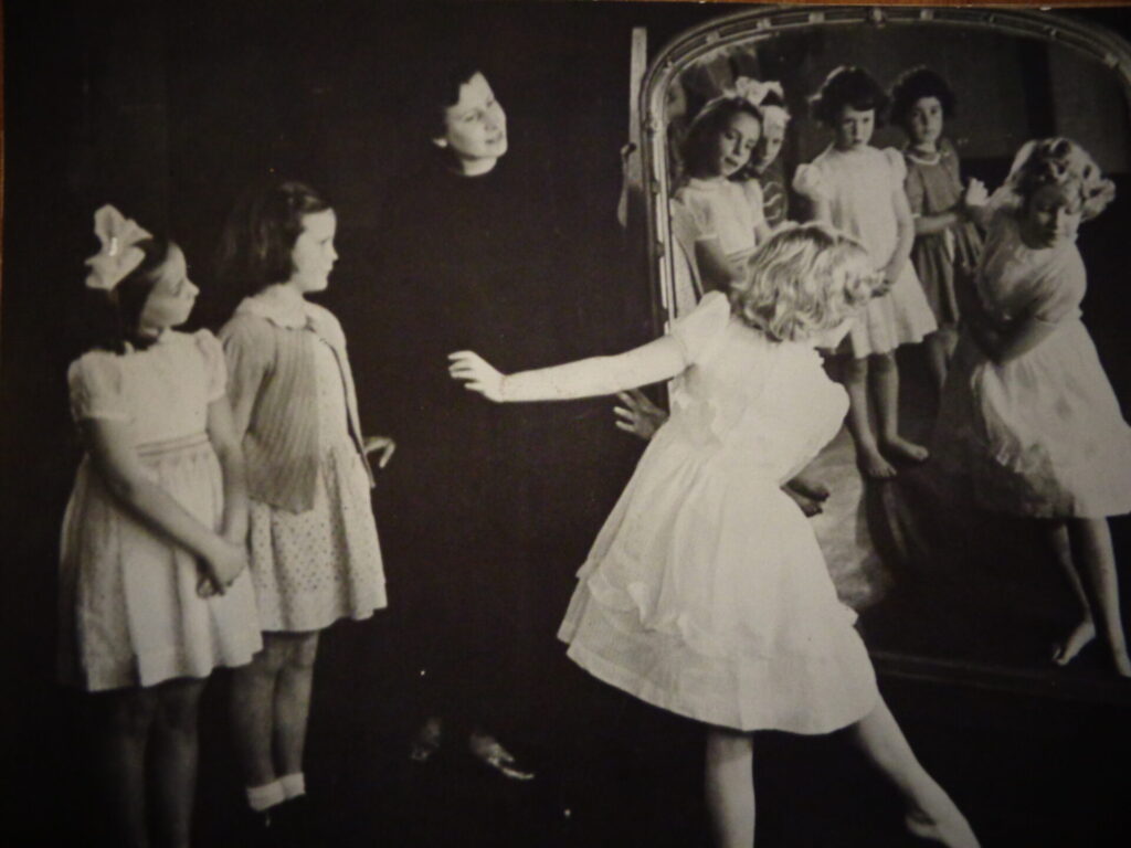 Girl practices dance move watched by two others and a teacher, reflected in a mirror. Black and white. Girls are wearing white and the teacher is in black.