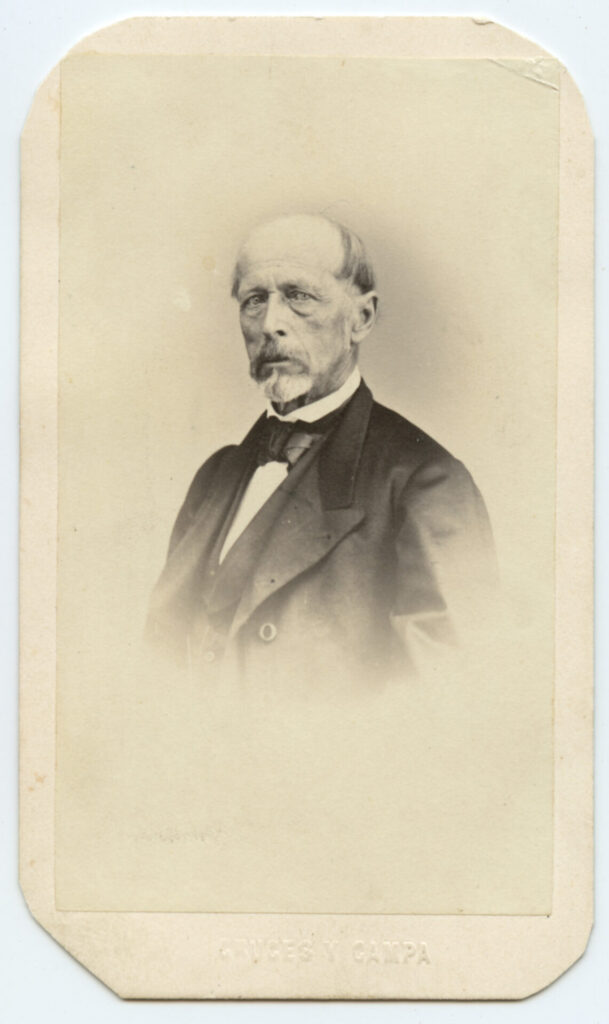 Black and white, daguerrotype image of man in a three-piece black suit with wide lapels and a bow tie. He is bearded and mustached and mostly bald, with a stern expression.