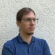 Nikolaus Hagen. Young man with glasses in semi-profile wearing a blue shirt with his long hair pulled back in a ponytail. He has a goatee but no moustache.
